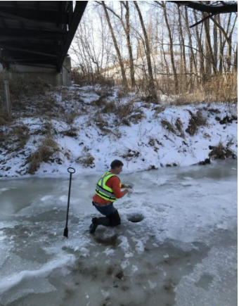 A person in a highlighter-yellow life vest kneels on a frozen creek with snowy banks