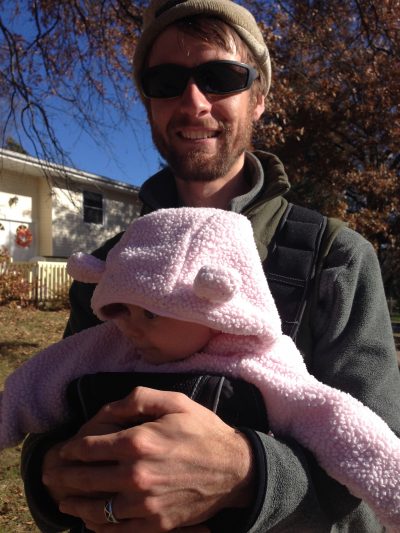 Greg smiles and holds a child in a pink bear suit in front of him.