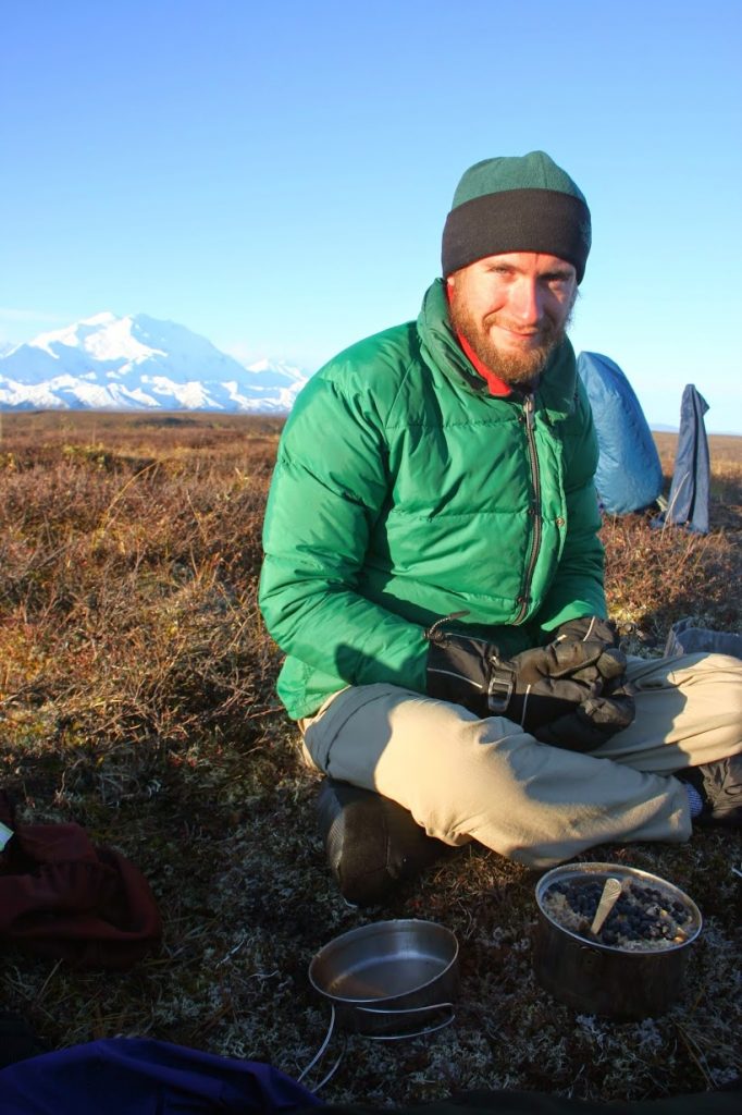 Greg sits in cold-weather gear with a mountain in the background